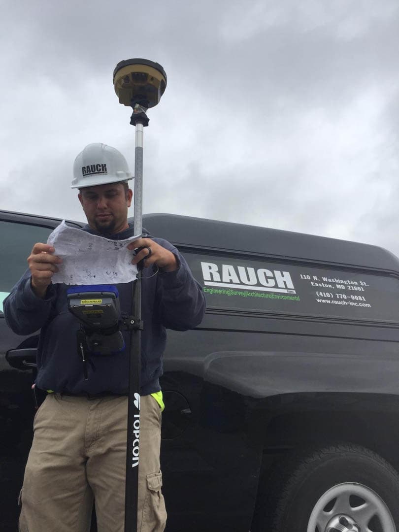 RAUCH survey technician in white hard hat reviewing printed field plans while standing beside a Topcon GPS rover rod, with a branded RAUCH van nearby