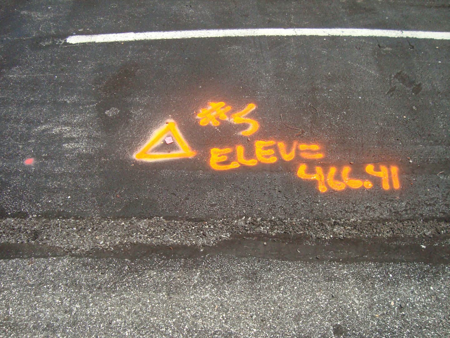 Close-up of orange spray-painted survey benchmark on dark asphalt pavement showing a triangle symbol with elevation notation