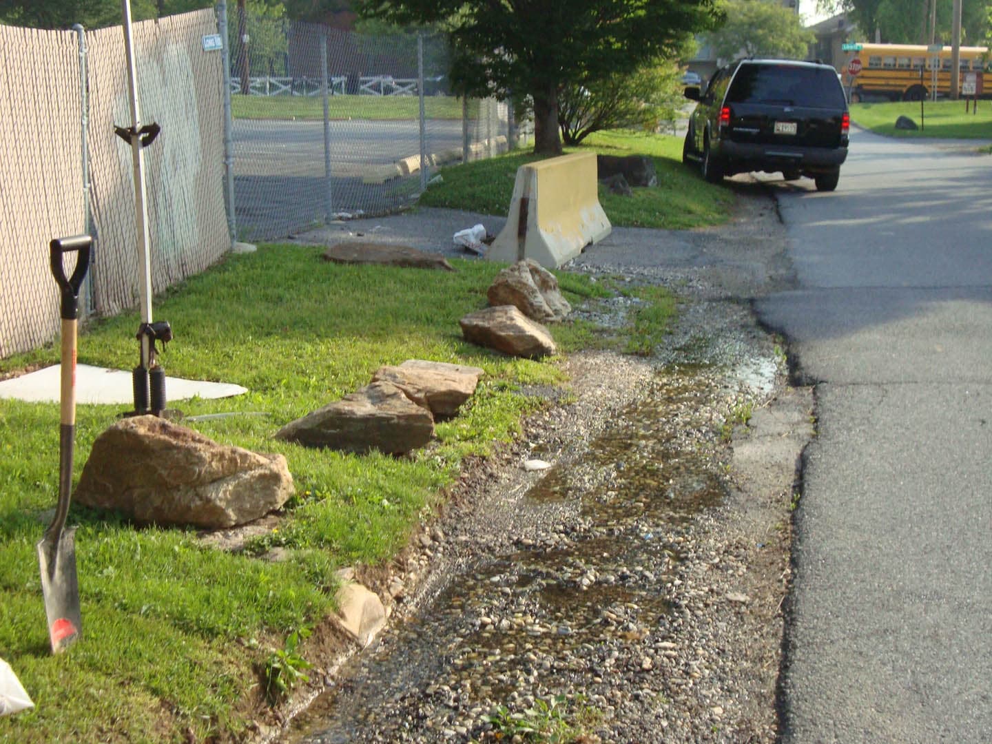 Roadside survey scene showing survey stakes and shovels planted along a gravel drainage swale beside an asphalt road with large boulders