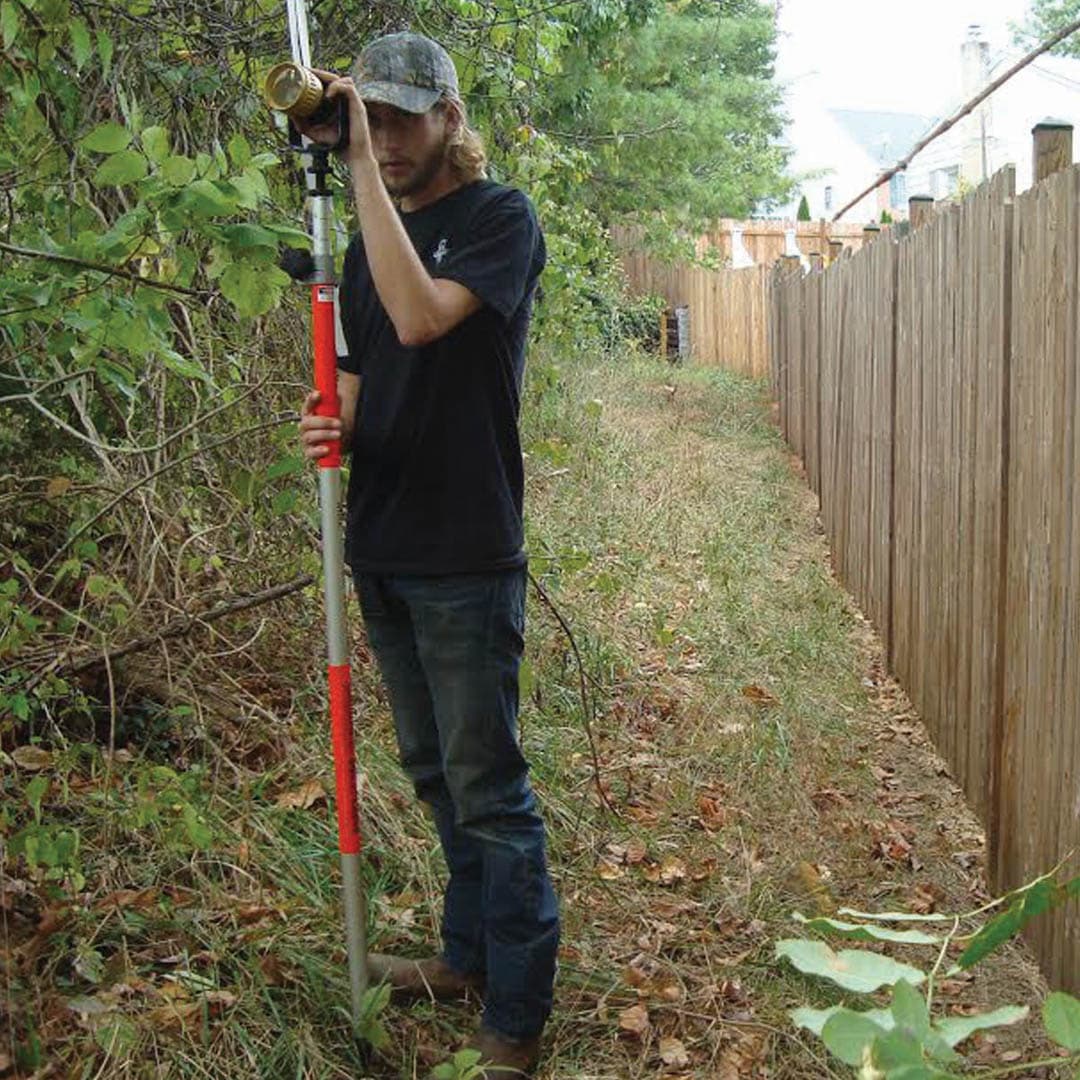Survey technician sighting through an optical level instrument along a narrow wooded corridor between a privacy fence and dense brush