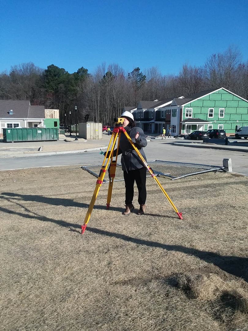 Survey technician in hard hat operating a total station on a yellow tripod at a residential construction site with newly framed townhomes in the background