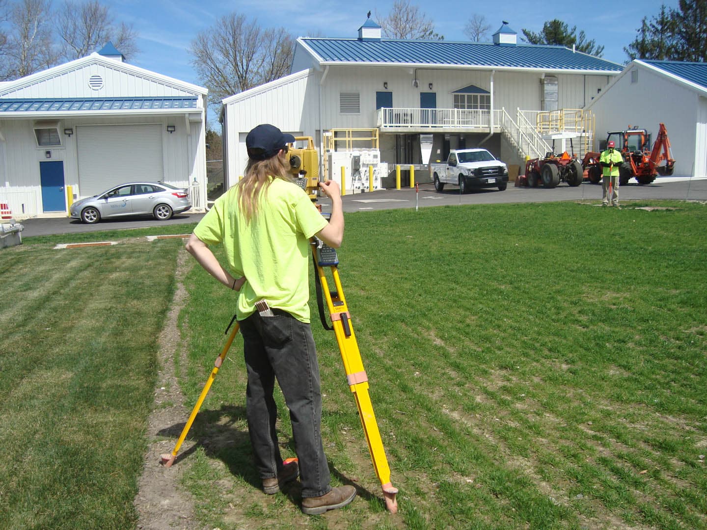 Survey technician operating a total station on a tripod in front of a white water treatment facility building with blue metal roof