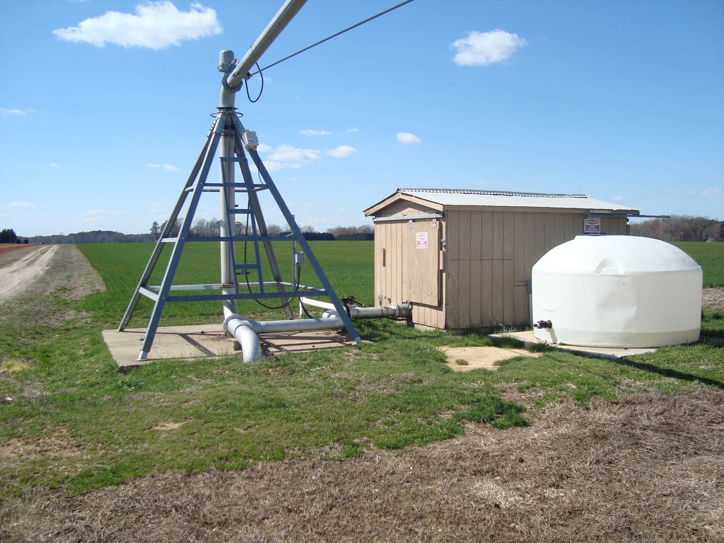 Rural agricultural pump station with a metal tripod-mounted pump, a small wooden utility shed, and a white dome-shaped storage tank beside a flat farm field