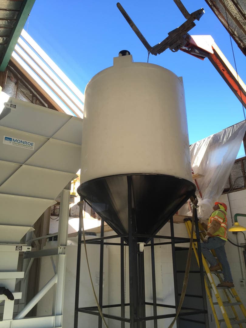 Tall white conical-bottom chemical feed tank on a steel stand being installed inside a water treatment facility