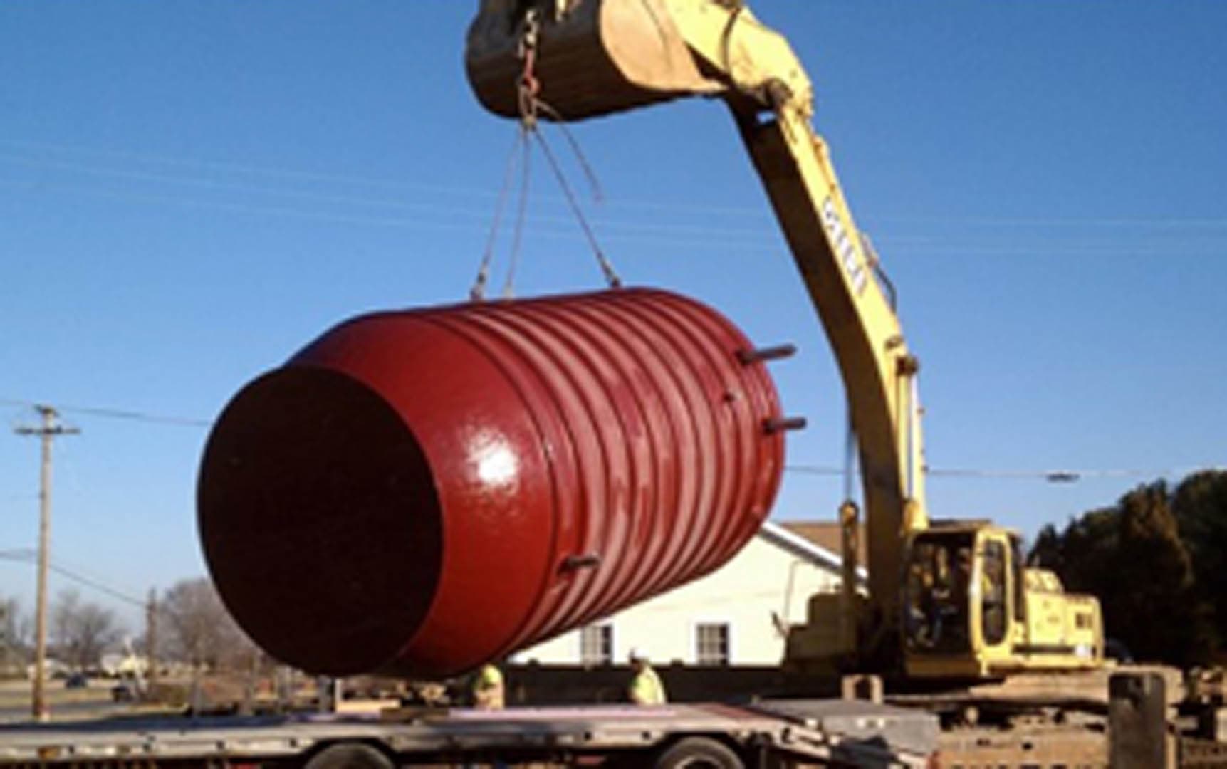 Excavator crane lifting a large corrugated red cylindrical tank horizontally off a flatbed trailer at a construction site