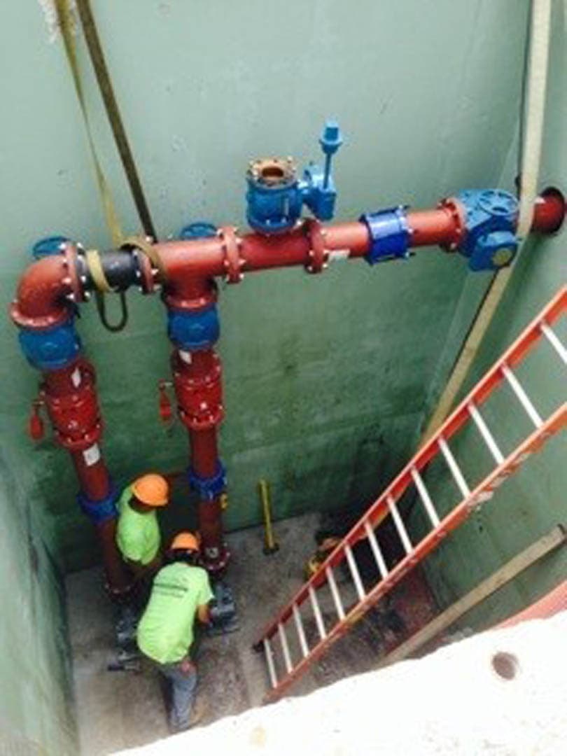 Workers inside a concrete pump station wet well installing red pressurized pipes with blue valves and fittings
