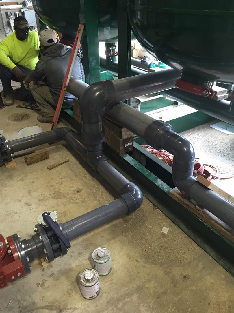Workers assembling a gray PVC pipe manifold connecting to large green pressure filter vessels on the floor of a water treatment facility