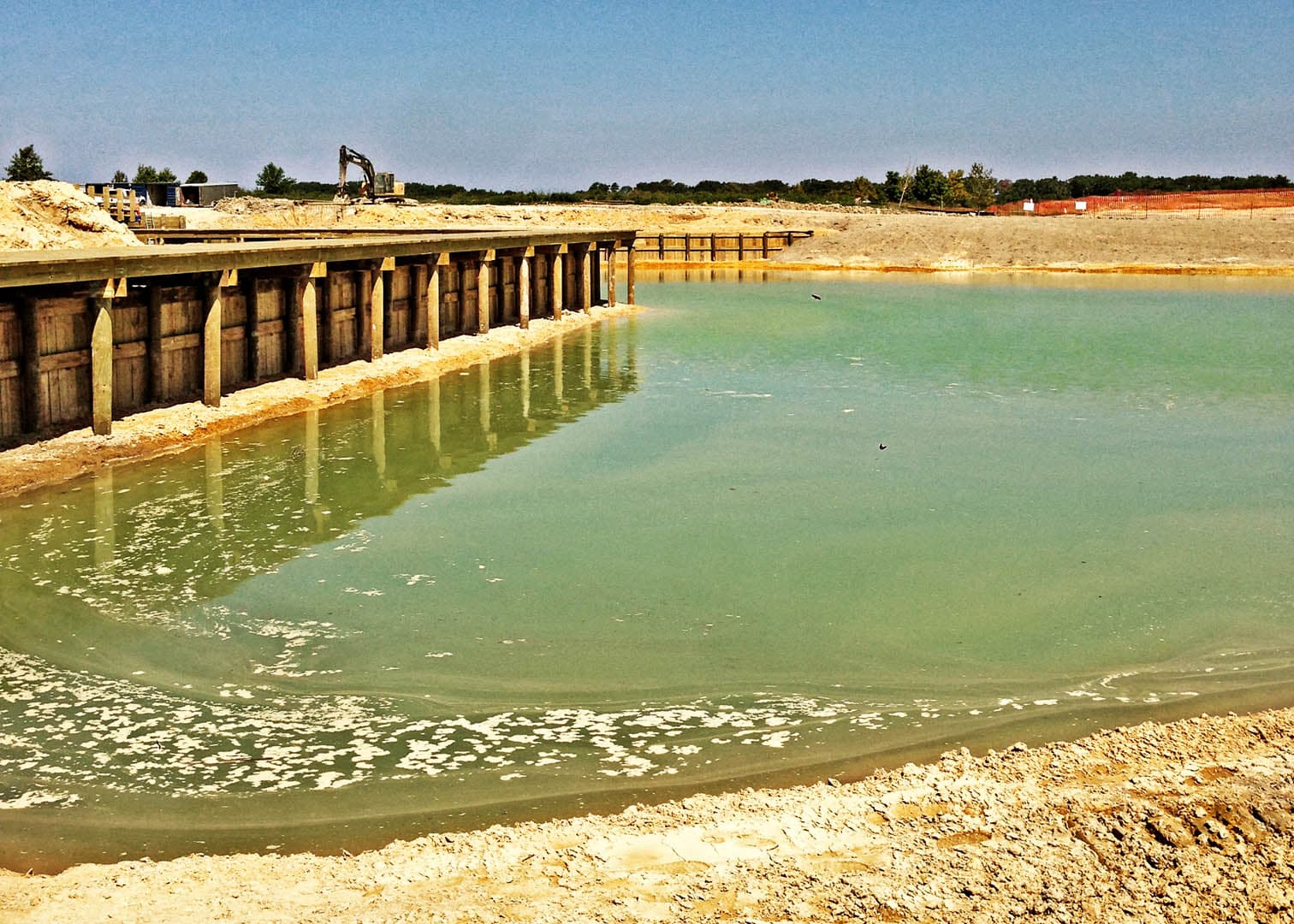 Large wastewater treatment lagoon under construction with a timber sheet pile bulkhead retaining wall and an excavator in the background