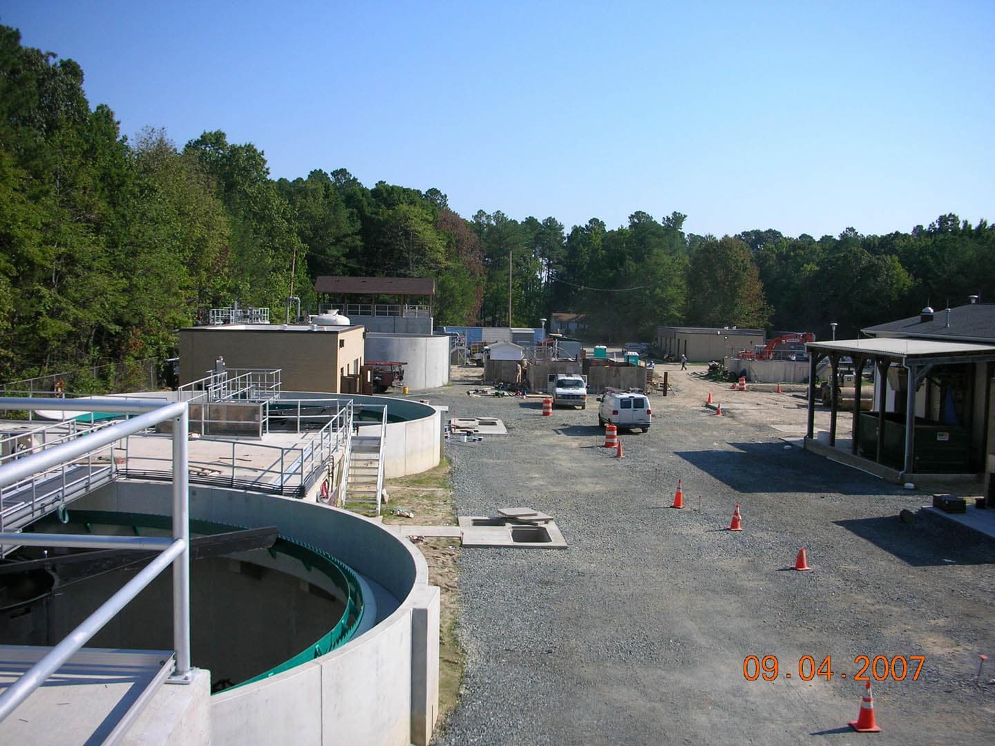 Aerial overview of a wastewater treatment plant showing circular clarifier tanks, process buildings, gravel roads, and construction vehicles