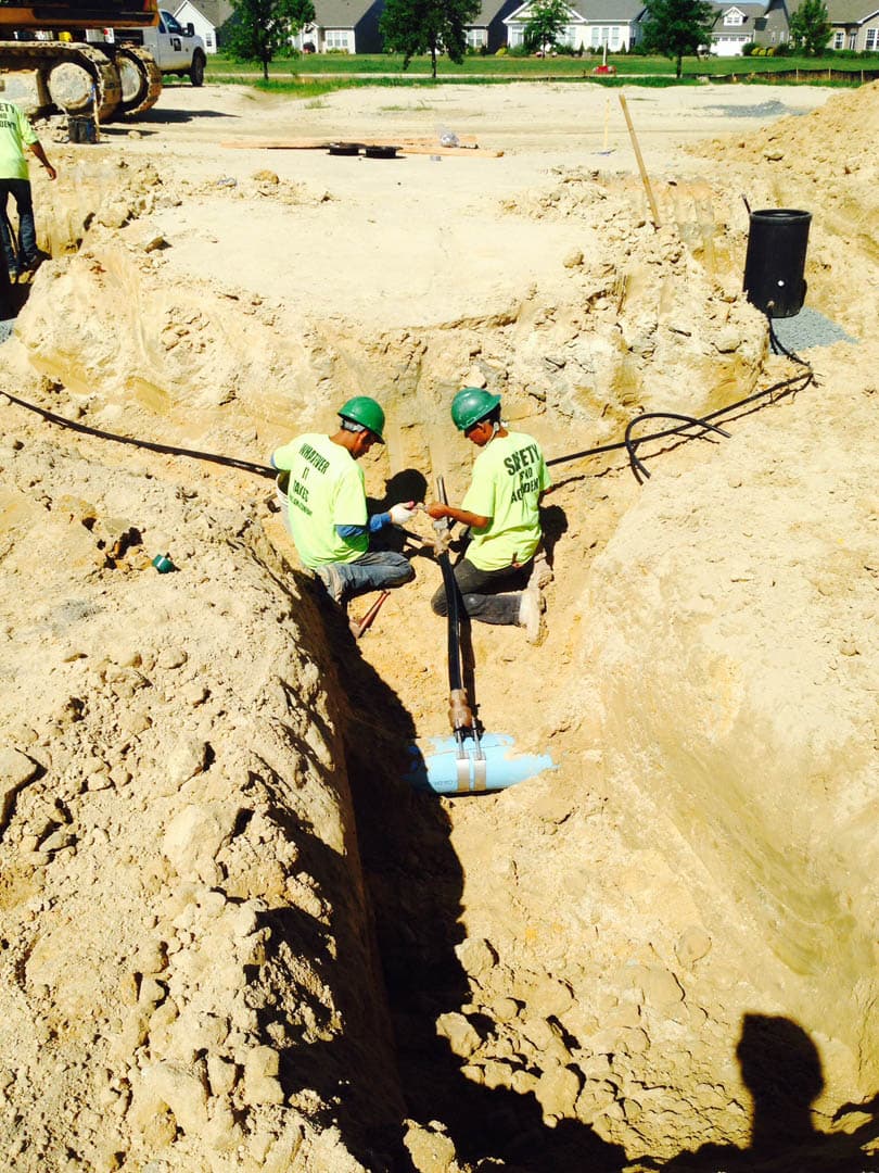 Two workers in safety vests kneeling in a sandy trench connecting a water service pipe to a larger water main in a residential development
