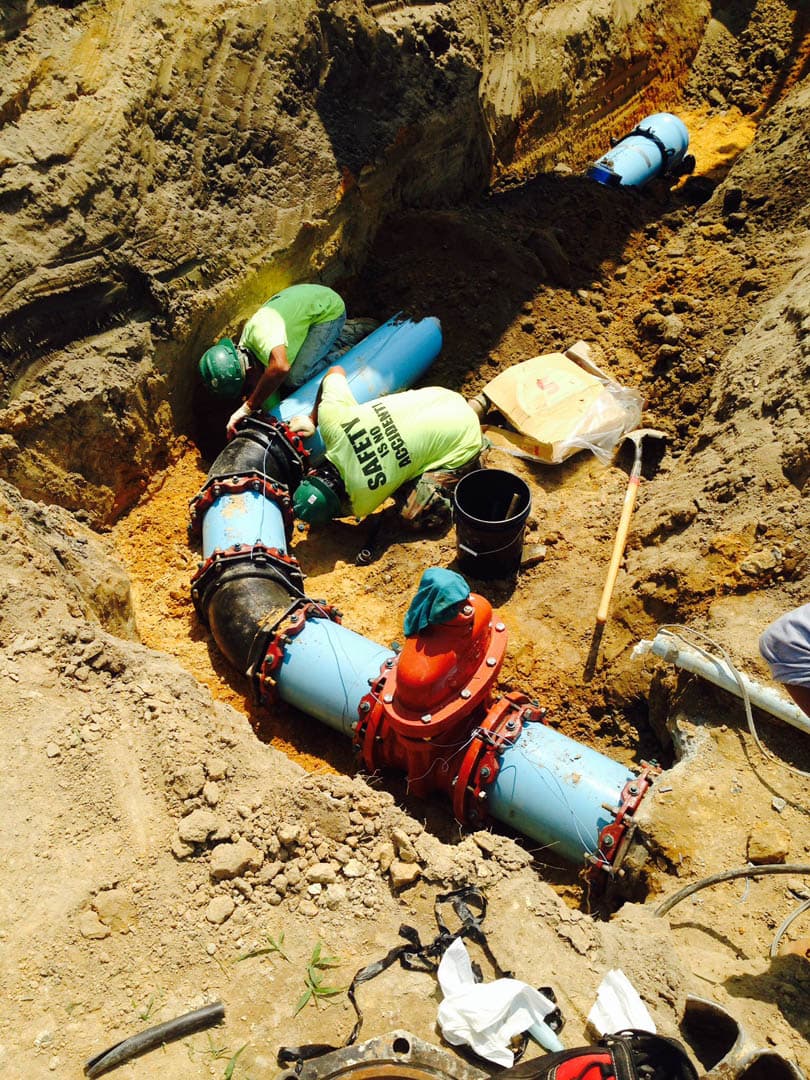 Two workers in safety vests assembling large blue water main pipes with red mechanical couplings in an open excavation trench