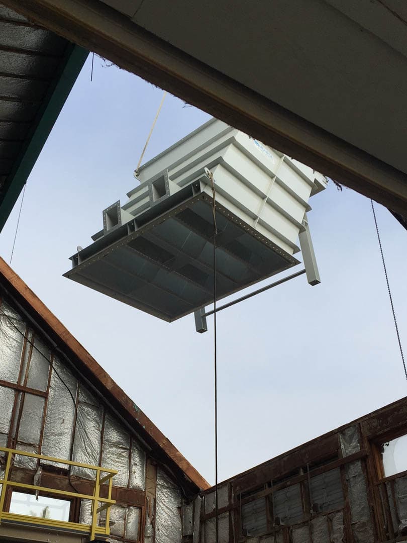 Large white metal water treatment equipment unit being lifted by crane through an open roof hatch into a treatment facility building
