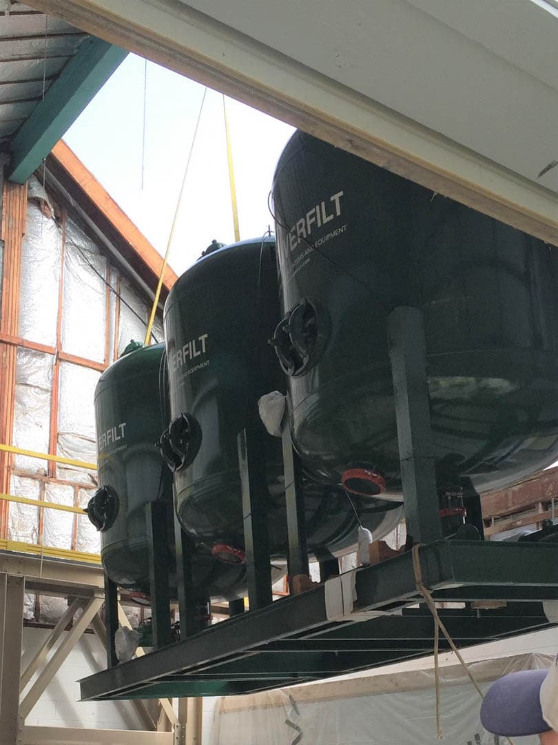 Three large dark green Everfilt pressure filter vessels being lowered through a roof opening into a water treatment building using lifting straps